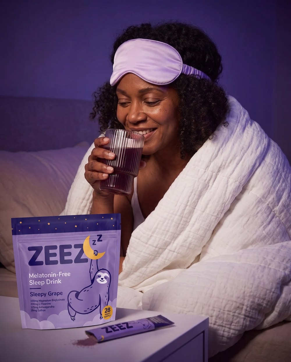A woman wrapped in a blanket holding a prepared Sleepy Grape drink beside ZEEZ packaging